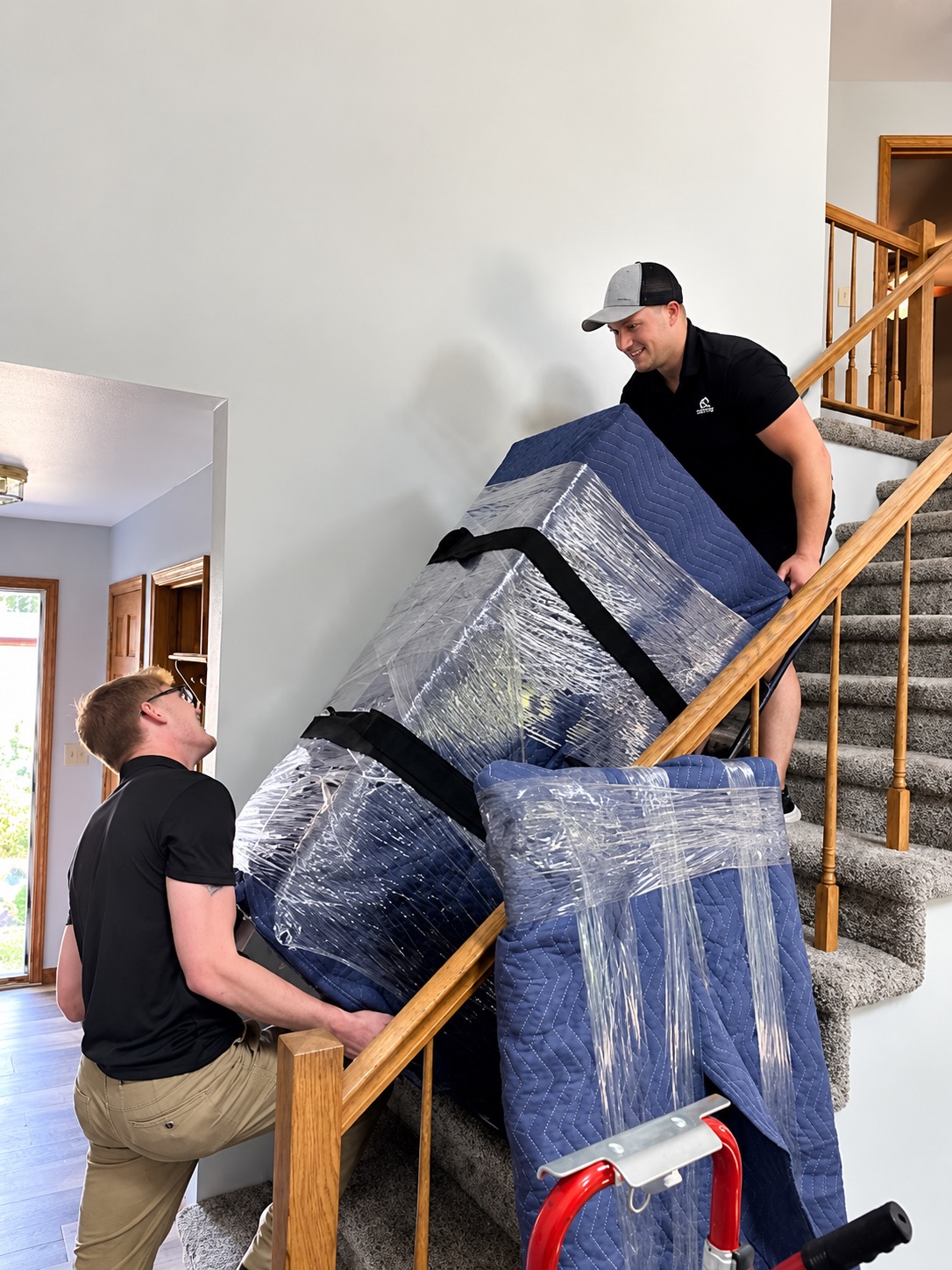 Two Peak Movers crew members carefully carrying a blanket-wrapped piece of furniture up a carpeted staircase